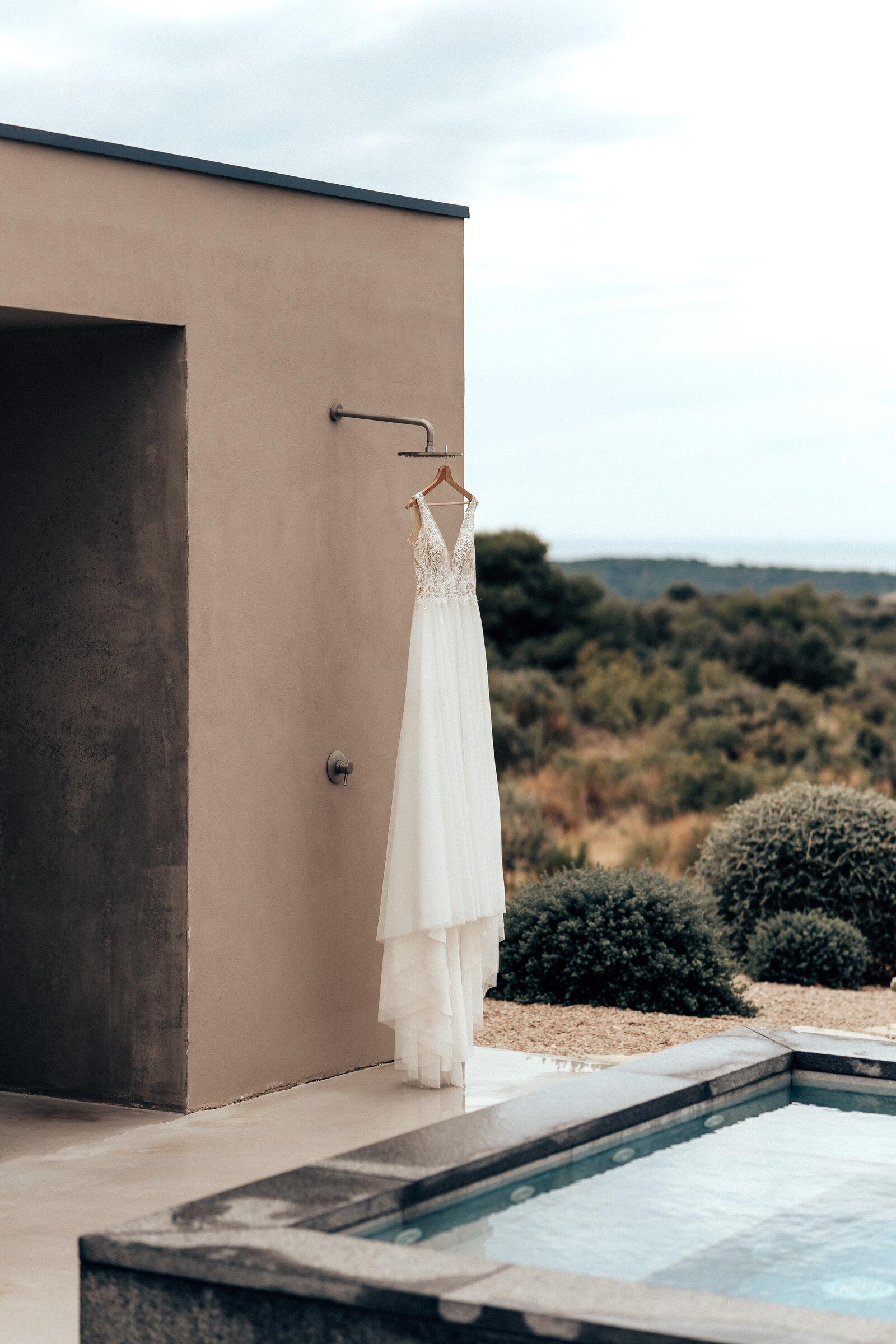 Brautkleid hängt an einer Außendusche neben dem Pool, dahinter weite Landschaft