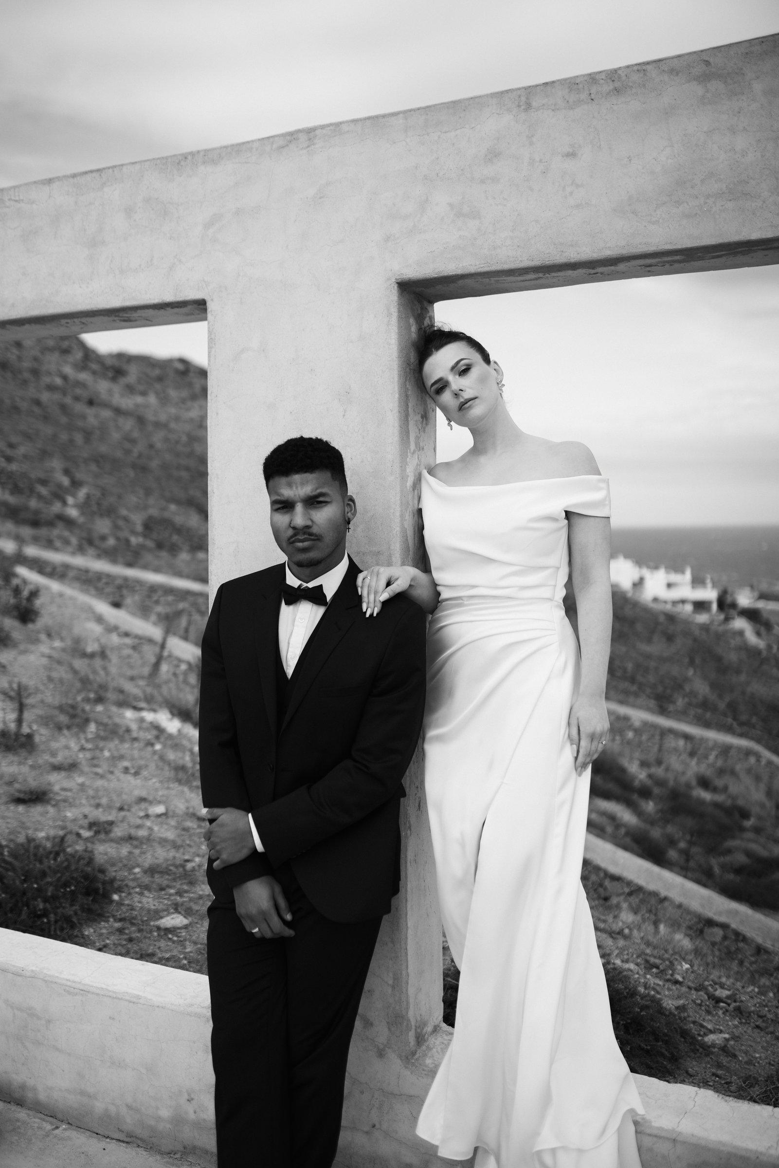 Braut und Bräutigam lehnen an einem Betonrahmen mit Blick auf Hügel und Meer auf Mykonos, Schwarzweiß